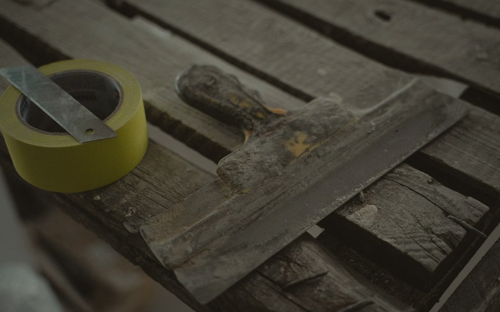 A worn putty knife and roll of yellow tape resting on gapped wooden floorboards